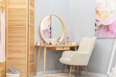 Mirror, dressing table with cosmetic products, armchair and folding screen indoors. Interior design