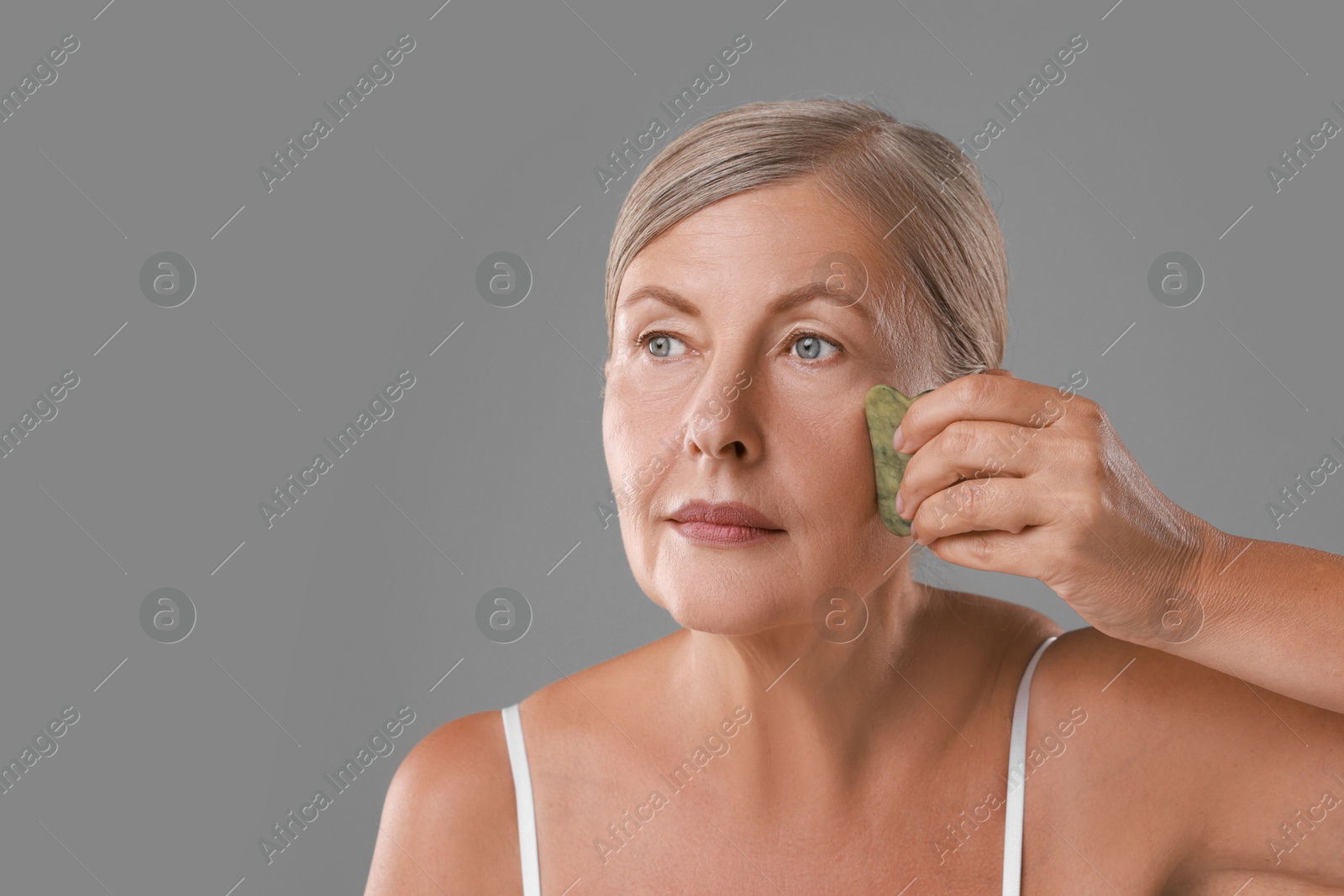 Photo of Beautiful woman doing facial massage with gua sha tool on grey background, space for text