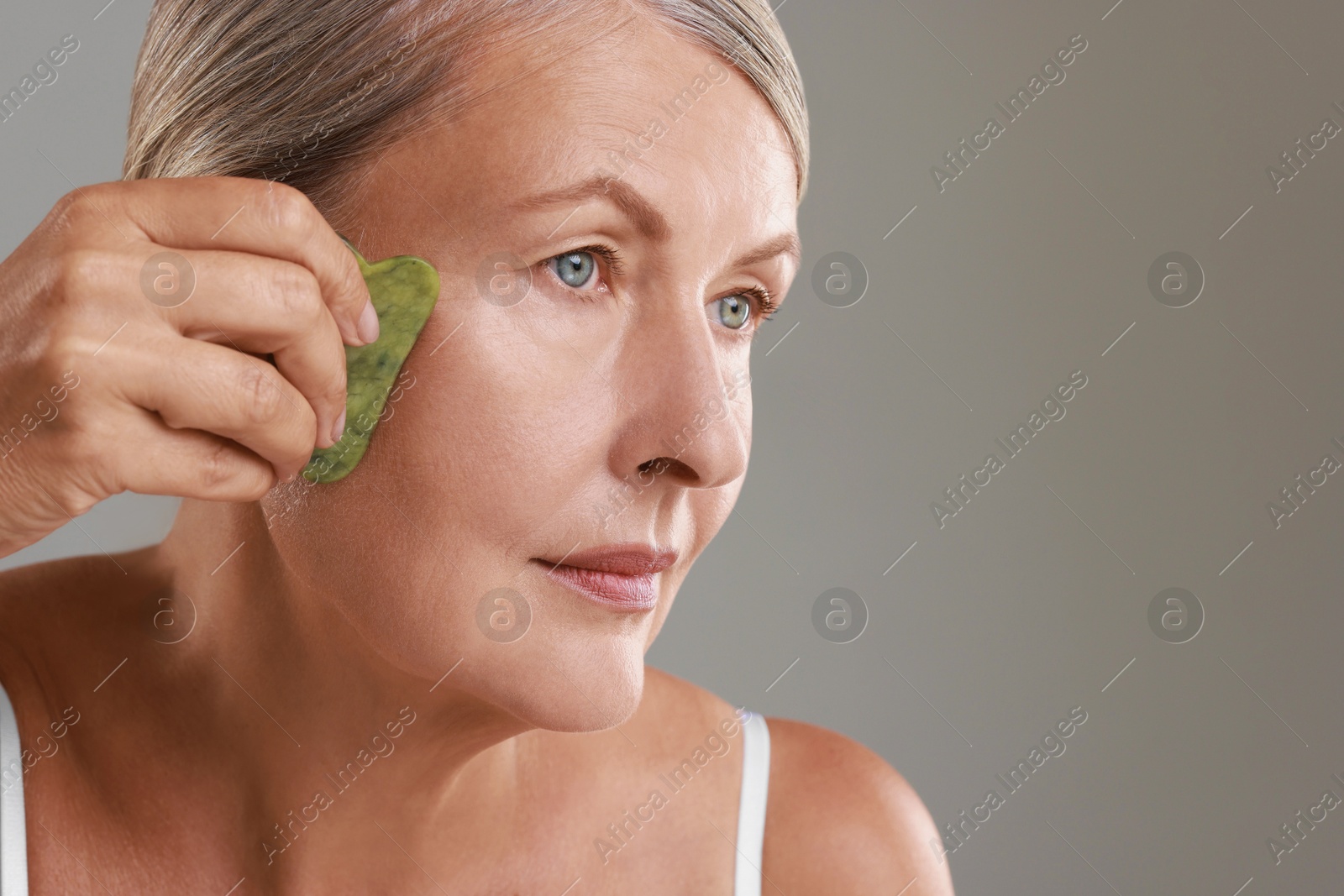 Photo of Beautiful woman doing facial massage with gua sha tool on grey background, closeup. Space for text