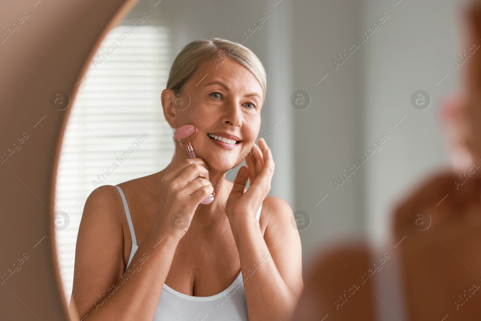 Photo of Beautiful woman doing facial massage with roller near mirror at home