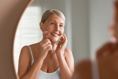 Beautiful woman doing facial massage with roller near mirror at home