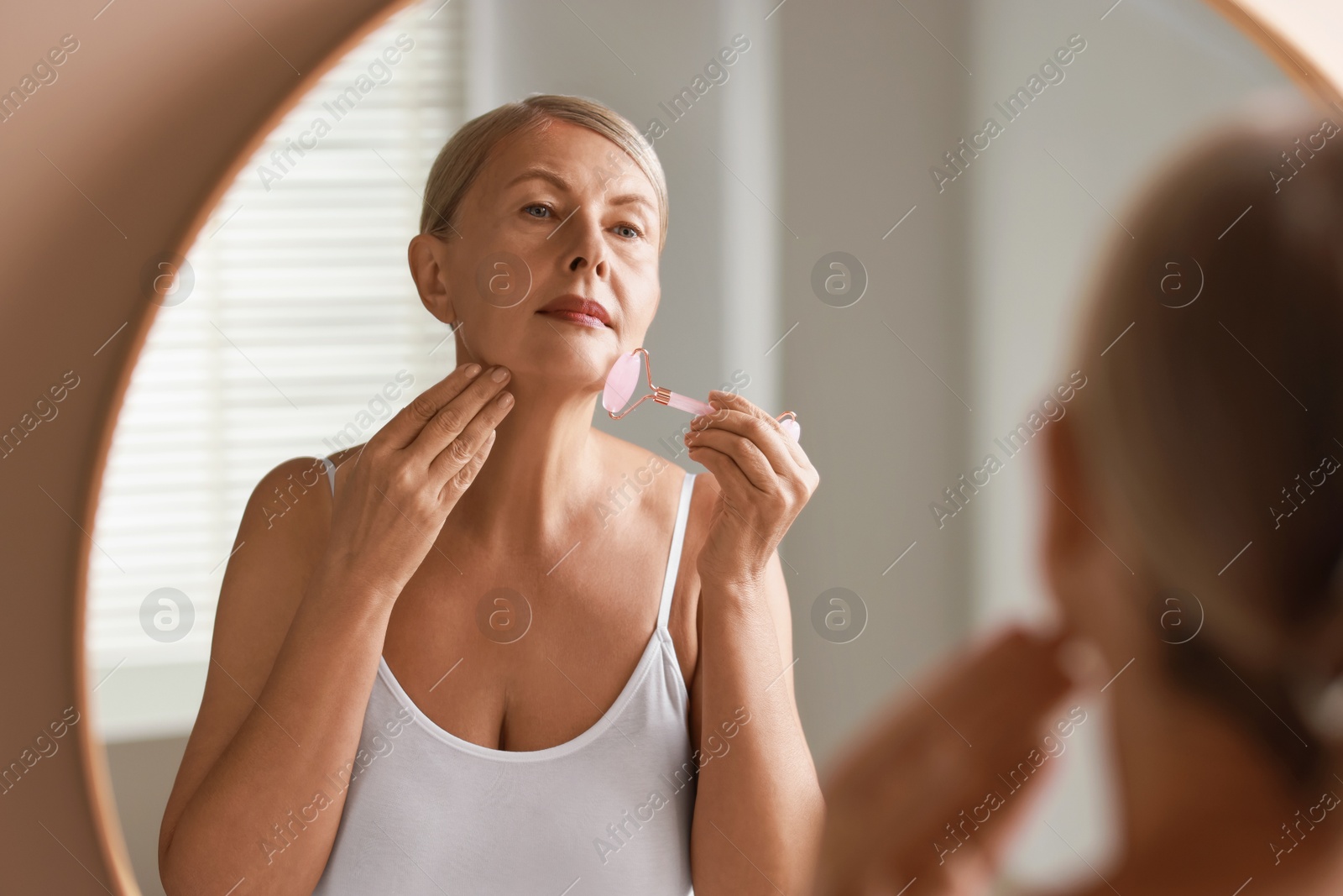 Photo of Beautiful woman doing facial massage with roller near mirror at home