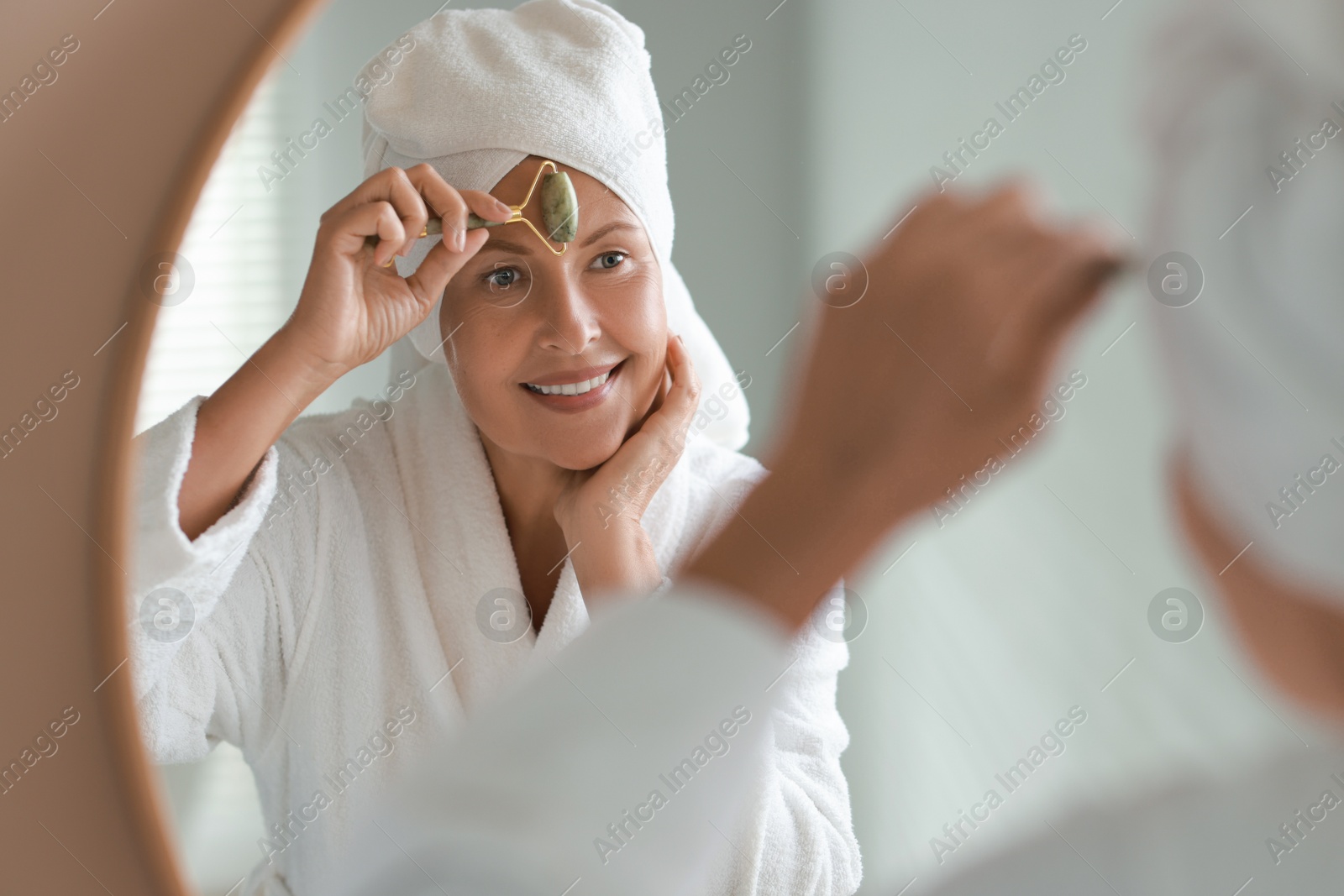 Photo of Beautiful woman doing facial massage with roller near mirror at home