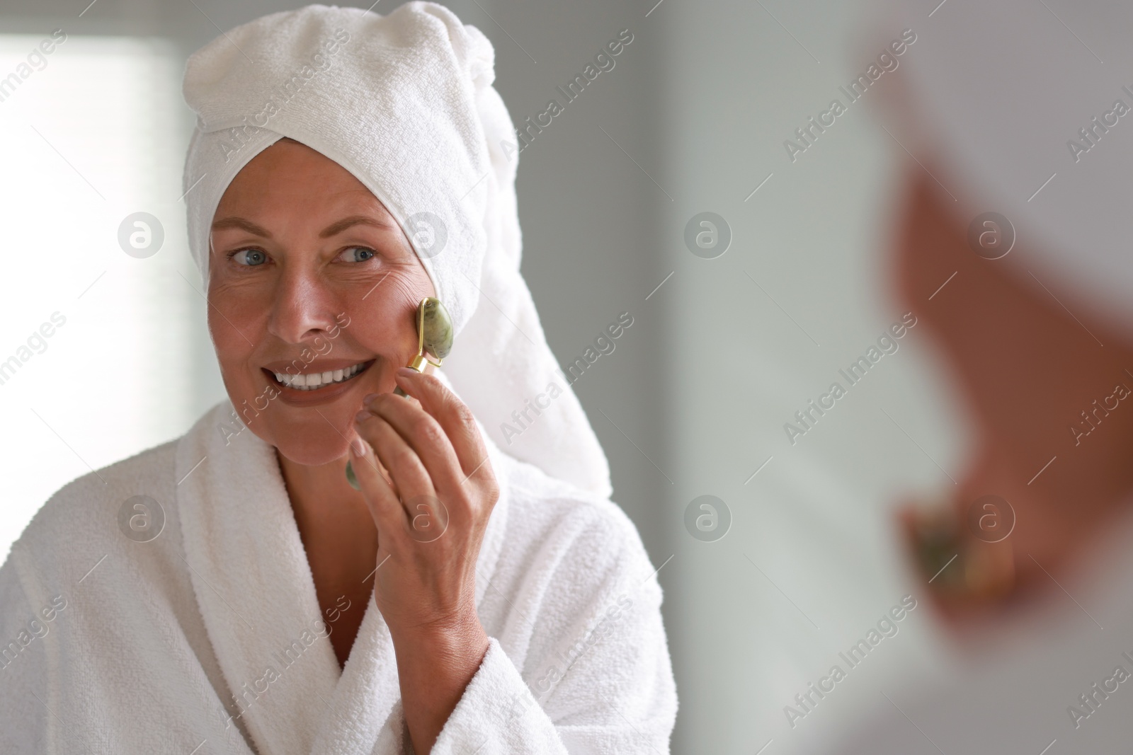 Photo of Beautiful woman doing facial massage with roller near mirror at home