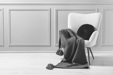 Image of Comfortable armchair with blanket and pillow near wall indoors, black and white