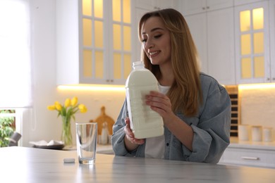 Photo of Young woman pouring milk from gallon bottle into glass at white marble table in kitchen