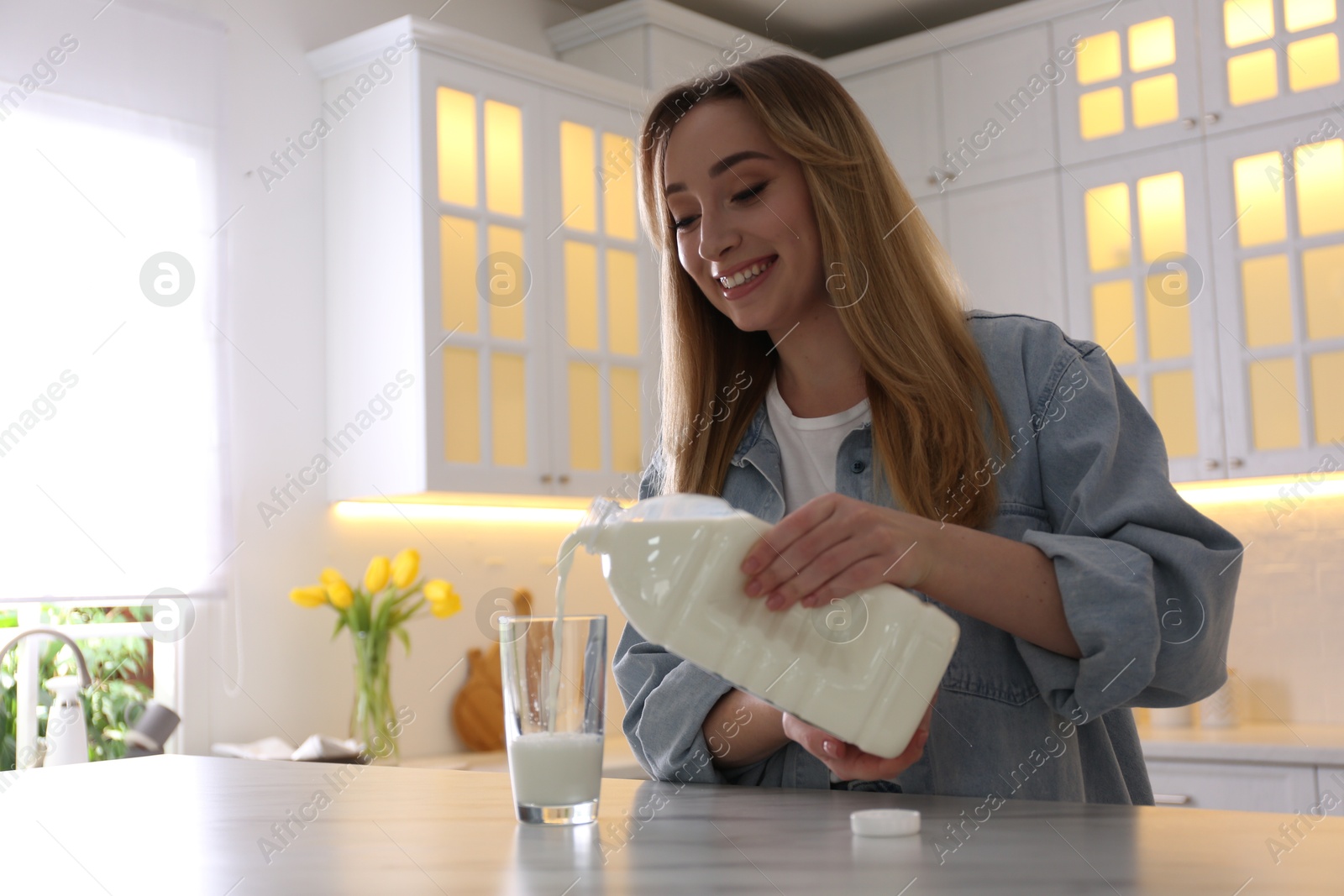 Photo of Young woman pouring milk from gallon bottle into glass at white marble table in kitchen