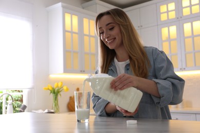 Young woman pouring milk from gallon bottle into glass at white marble table in kitchen