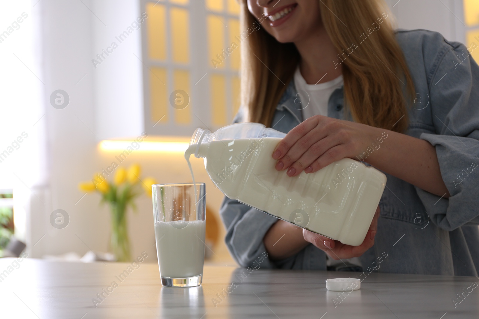 Photo of Young woman pouring milk from gallon bottle into glass at white marble table in kitchen, closeup