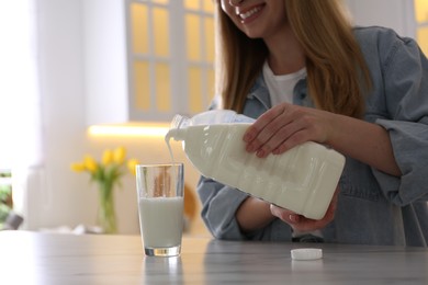 Young woman pouring milk from gallon bottle into glass at white marble table in kitchen, closeup