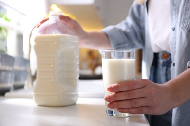 Photo of Young woman with gallon bottle of milk and glass at white countertop in kitchen, closeup