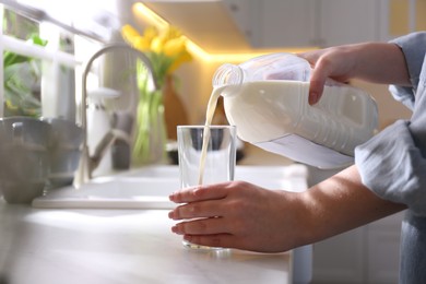 Photo of Young woman pouring milk from gallon bottle into glass at white countertop in kitchen, closeup