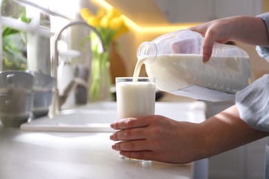 Photo of Young woman pouring milk from gallon bottle into glass at white countertop in kitchen, closeup