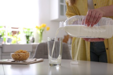 Young woman pouring milk from gallon bottle into glass at white table in kitchen, closeup