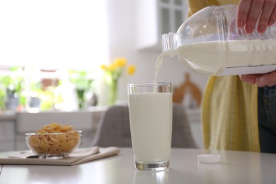 Young woman pouring milk from gallon bottle into glass at white table in kitchen, closeup