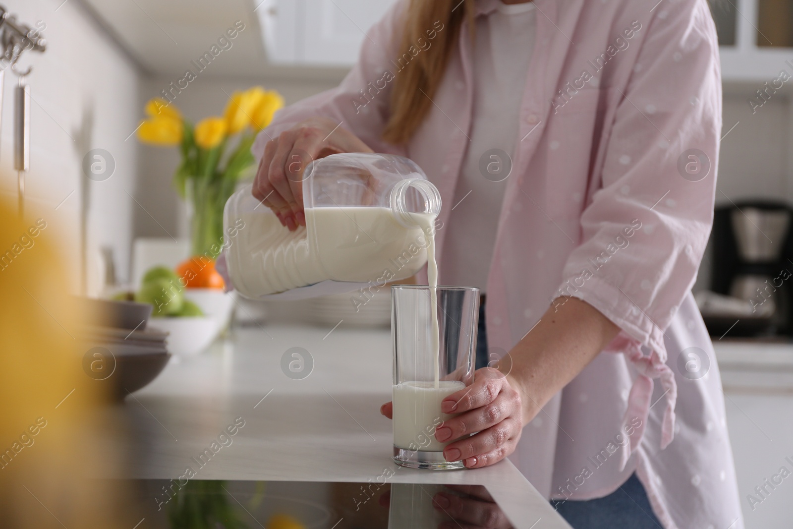 Photo of Young woman pouring milk from gallon bottle into glass at light countertop in kitchen, closeup