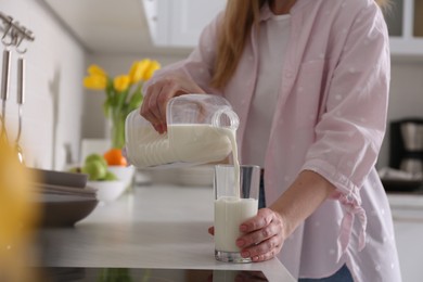 Young woman pouring milk from gallon bottle into glass at light countertop in kitchen, closeup