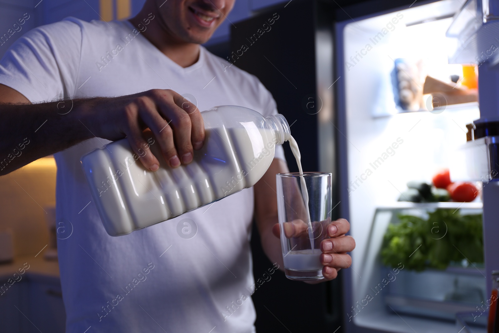 Photo of Man pouring milk from gallon bottle into glass near refrigerator in kitchen at night, closeup