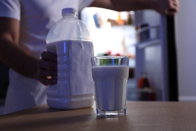 Man holding gallon bottle of milk and glass on wooden table in kitchen at night, closeup