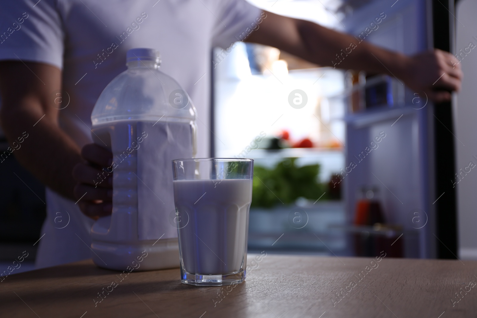 Photo of Man holding gallon bottle of milk and glass on wooden table in kitchen at night, closeup