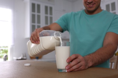 Man pouring milk from gallon bottle into glass at wooden table in kitchen, closeup