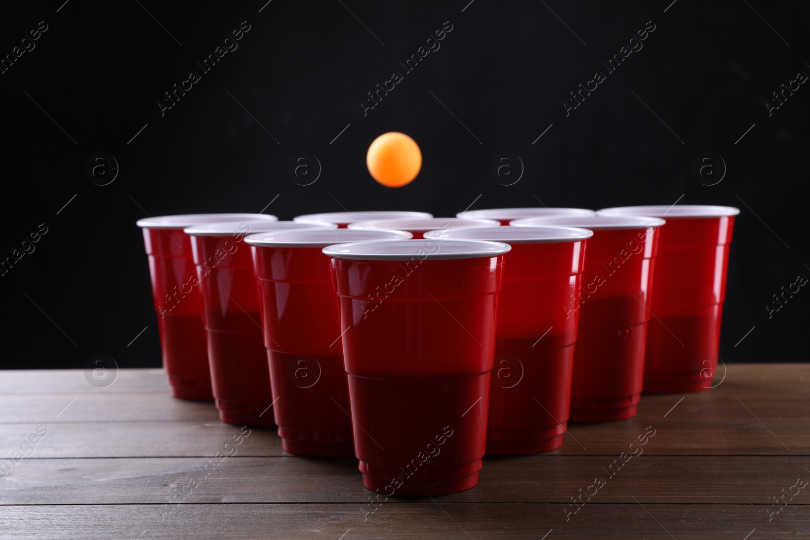 Photo of Plastic cups and ball for beer pong on wooden table against black background