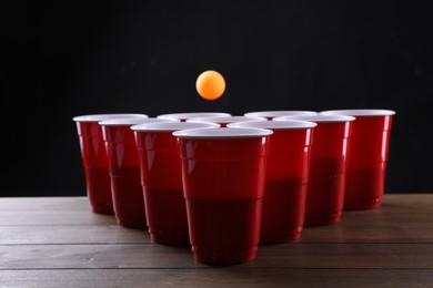 Photo of Plastic cups and ball for beer pong on wooden table against black background