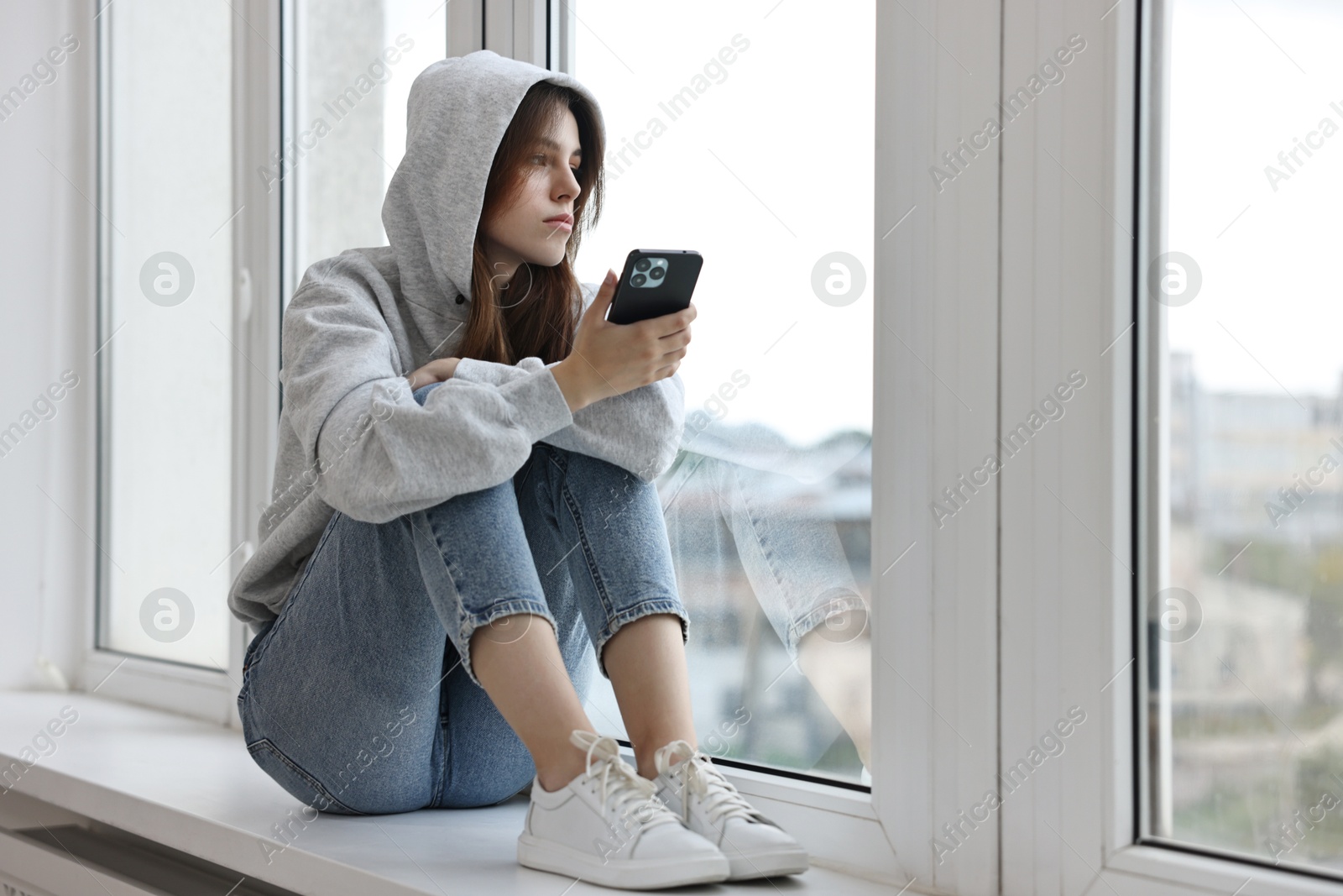 Photo of Loneliness concept. Sad teenage girl using smartphone on windowsill at home