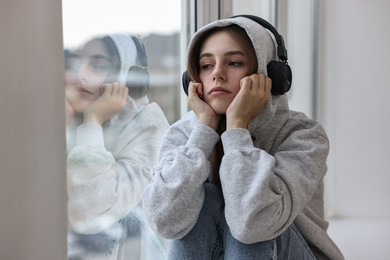 Loneliness concept. Sad teenage girl in headphones listening to music near window at home