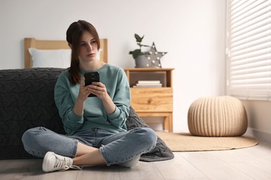 Loneliness concept. Sad teenage girl using smartphone on floor at home, space for text