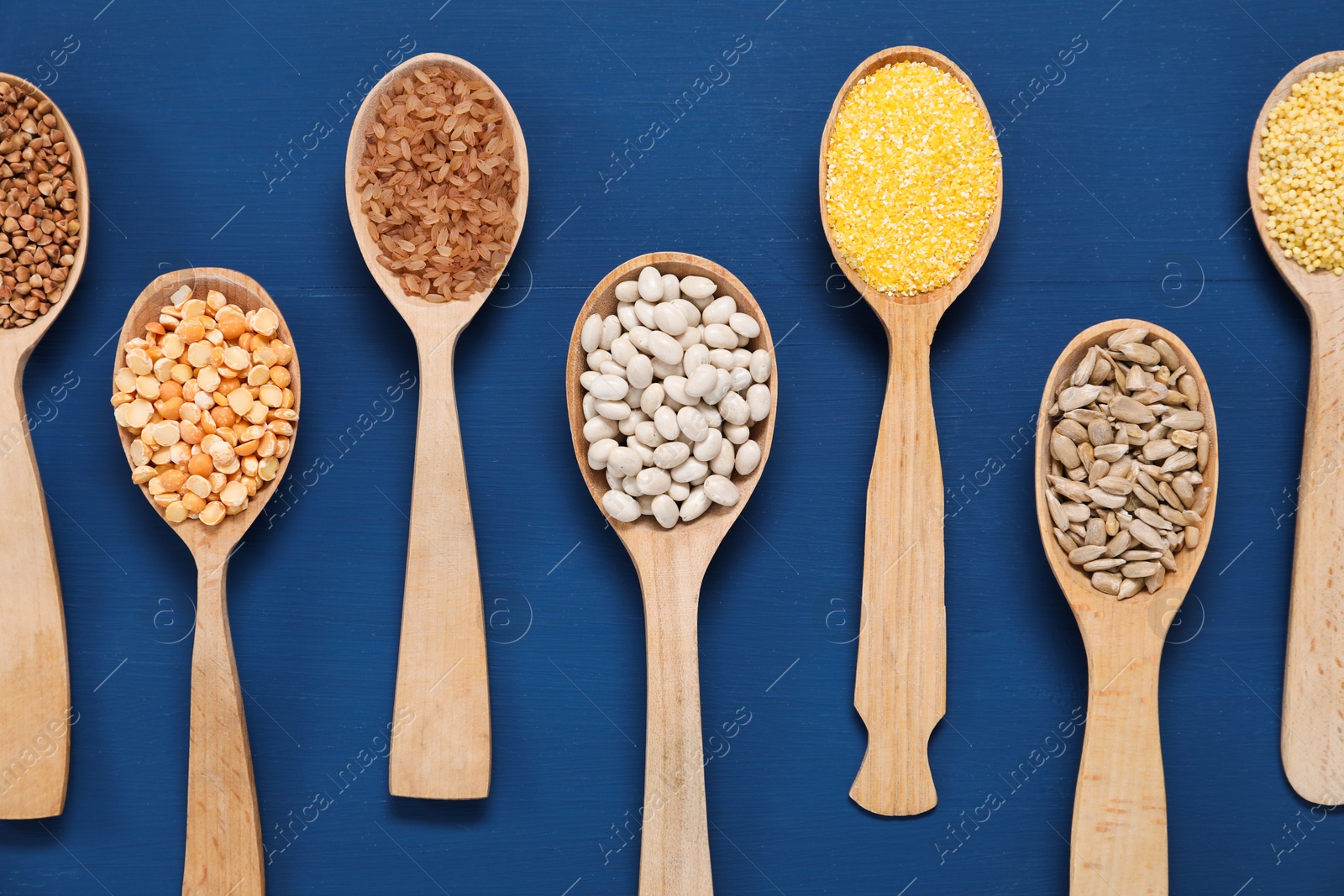 Photo of Different types of cereals, seeds and legumes in spoons on blue wooden table, flat lay