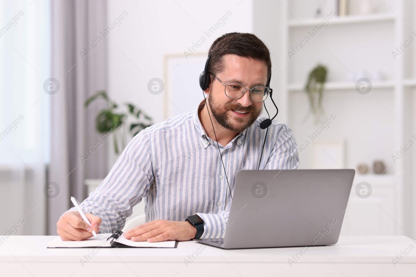 Photo of Interpreter in headset taking notes while having video chat via laptop at white table indoors