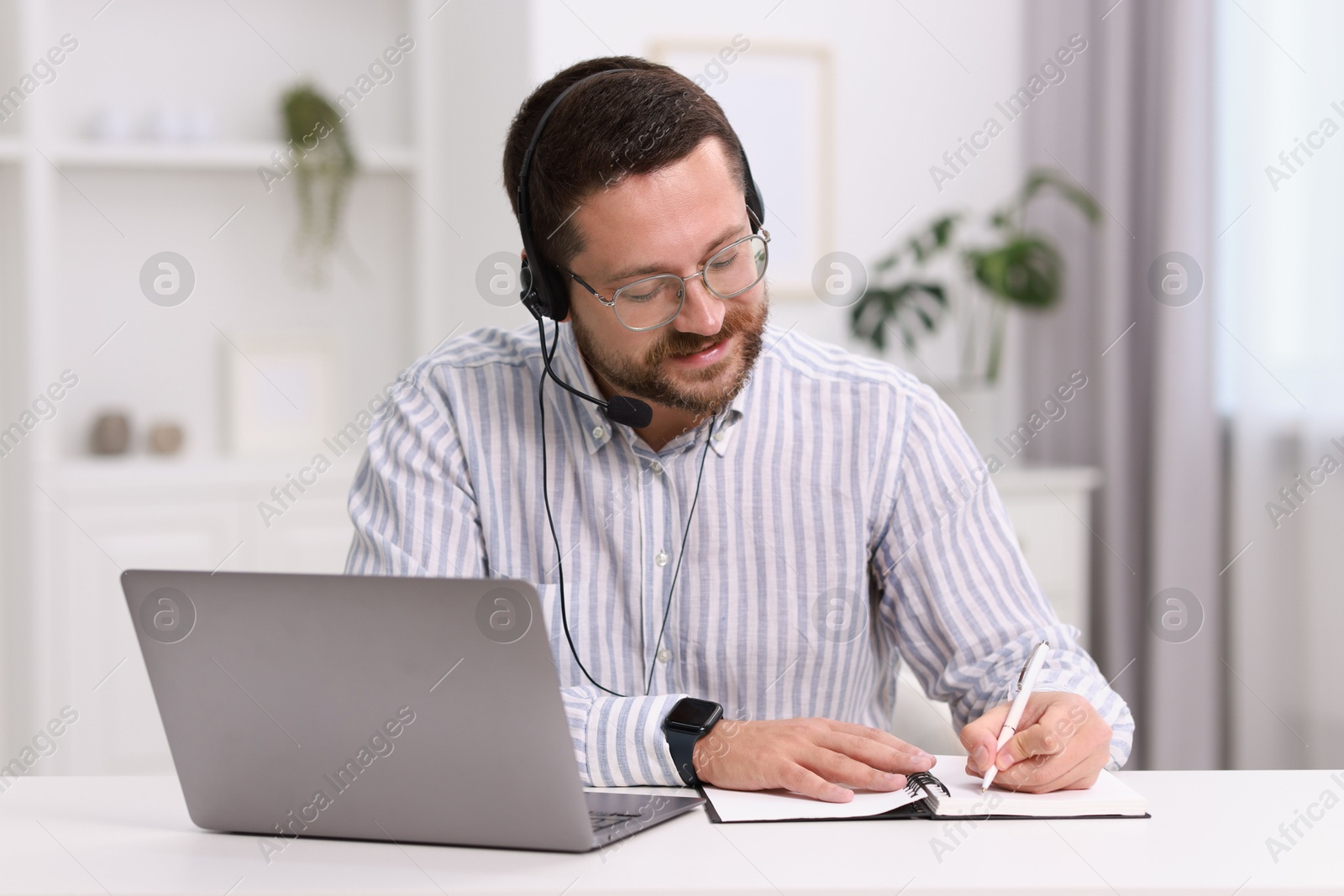 Photo of Interpreter in headset taking notes while having video chat via laptop at white table indoors