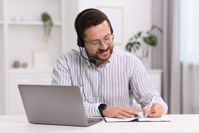 Interpreter in headset taking notes while having video chat via laptop at white table indoors