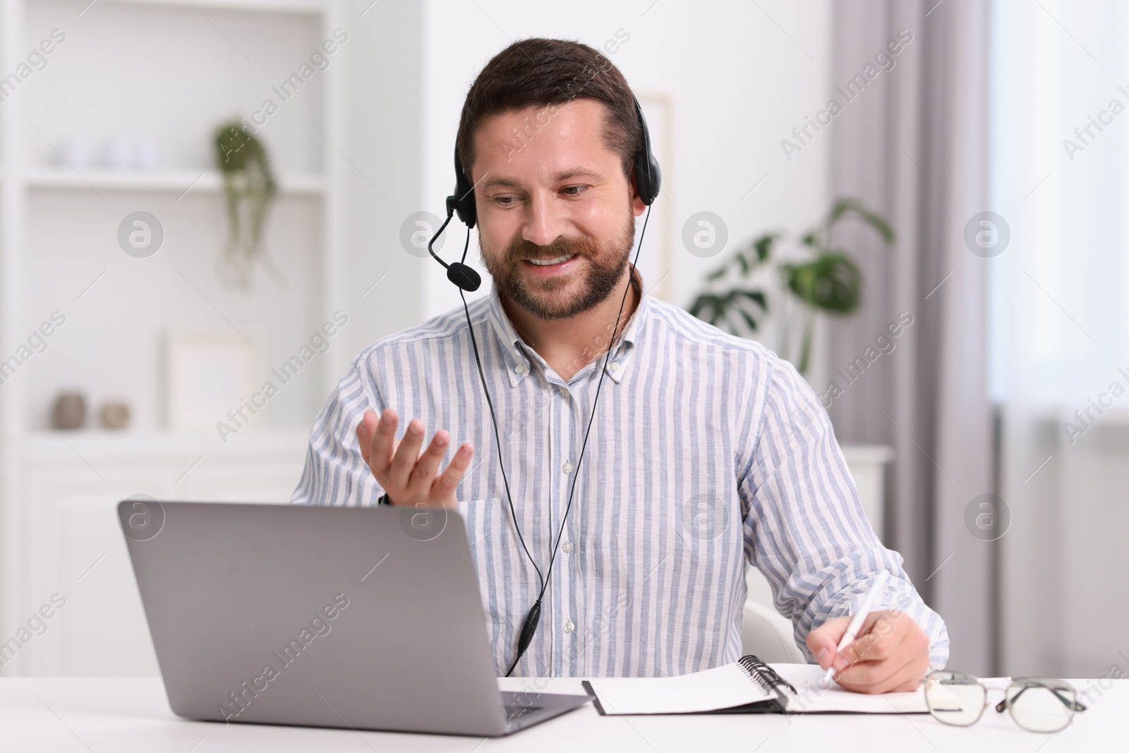 Photo of Interpreter in headset taking notes while having video chat via laptop at white table indoors