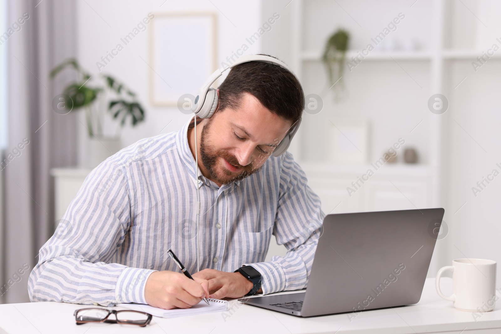 Photo of Interpreter in headphones taking notes while having video chat via laptop at white table indoors