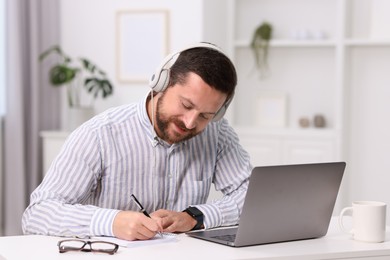 Photo of Interpreter in headphones taking notes while having video chat via laptop at white table indoors