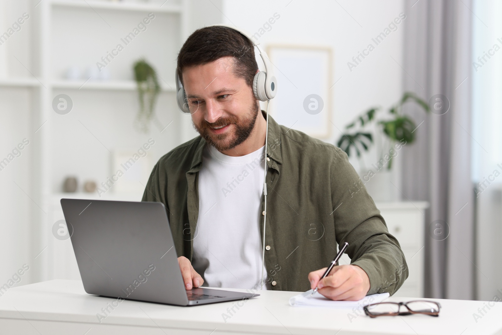 Photo of Interpreter in headphones taking notes while having video chat via laptop at white table indoors