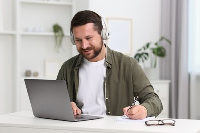 Interpreter in headphones taking notes while having video chat via laptop at white table indoors