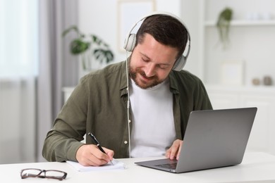 Photo of Interpreter in headphones taking notes while having video chat via laptop at white table indoors