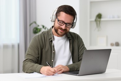 Photo of Interpreter in headphones taking notes while having video chat via laptop at white table indoors