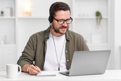 Interpreter in headset taking notes while having video chat via laptop at white table indoors