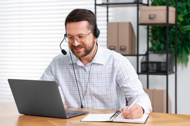 Interpreter in headset taking notes while having video chat via laptop at wooden table indoors