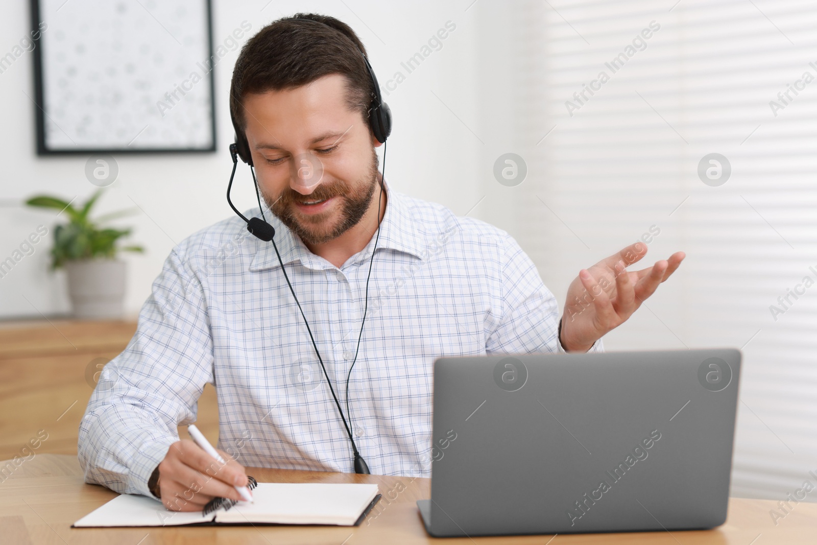 Photo of Interpreter in headset taking notes while having video chat via laptop at wooden table indoors