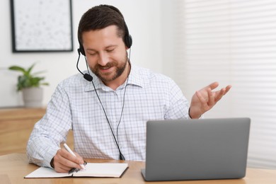 Interpreter in headset taking notes while having video chat via laptop at wooden table indoors