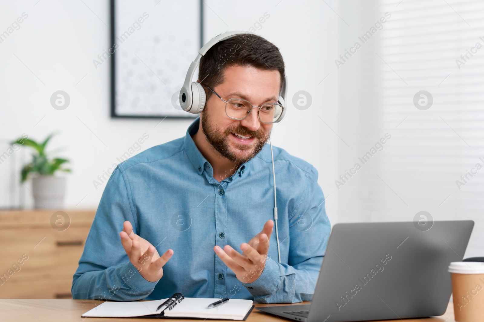 Photo of Interpreter in headphones having video chat via laptop at wooden table indoors