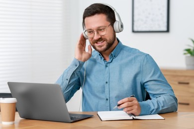 Interpreter in headphones taking notes while having video chat via laptop at wooden table indoors