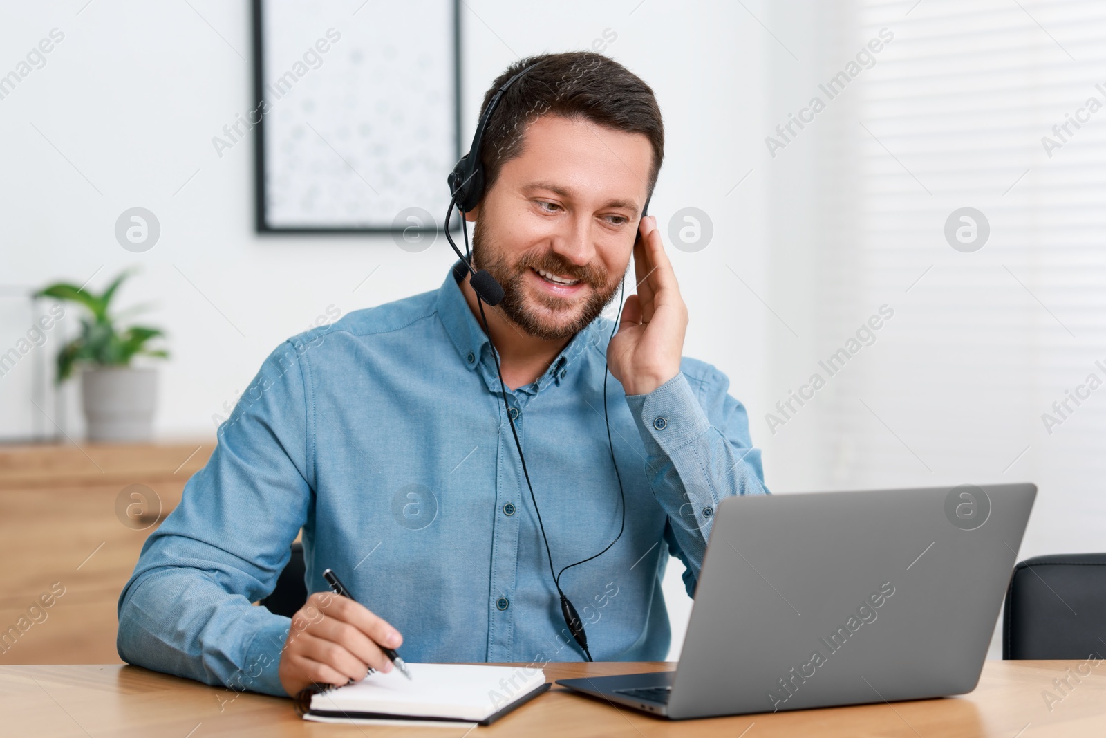 Photo of Interpreter in headset taking notes while having video chat via laptop at wooden table indoors