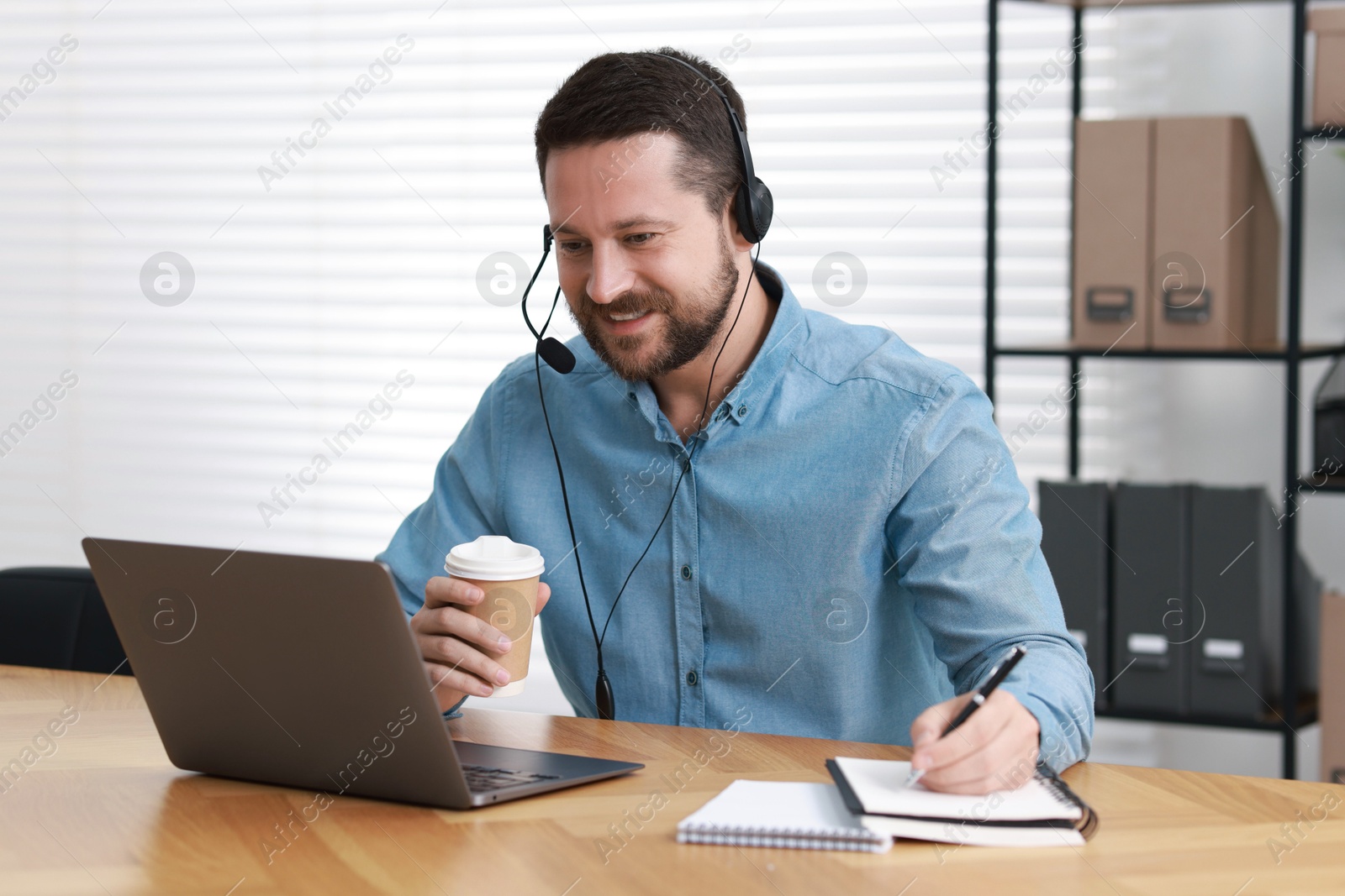 Photo of Interpreter in headset taking notes while having video chat via laptop at wooden table indoors