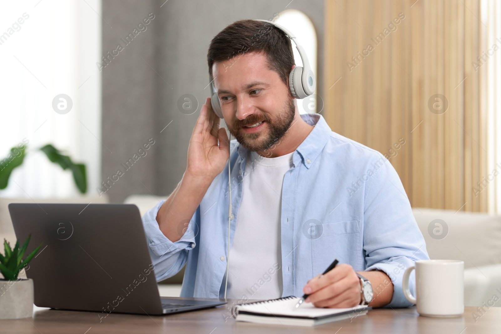 Photo of Interpreter in headphones taking notes while having video chat via laptop at wooden table indoors